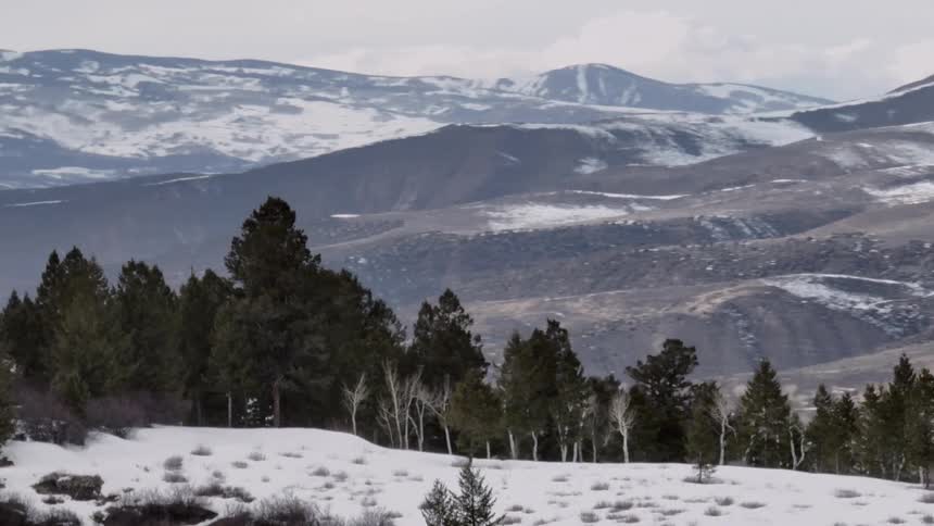 A scenic landscape shot captures a snow covered foreground leading to rolling hills and mountains in the distance with patches of snow and bare earth creating a textured pattern across the terrain