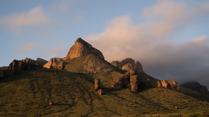 A rugged mountain peak its rocky surface illuminated by the warm light of sunrise or sunset stands prominently against a backdrop of a blue sky with soft pastel colored clouds