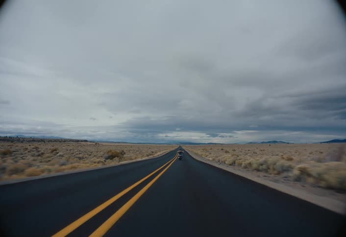 A lone motorcycle travels down a straight double yellowlined highway stretching across a desolate arid landscape under a heavy sky