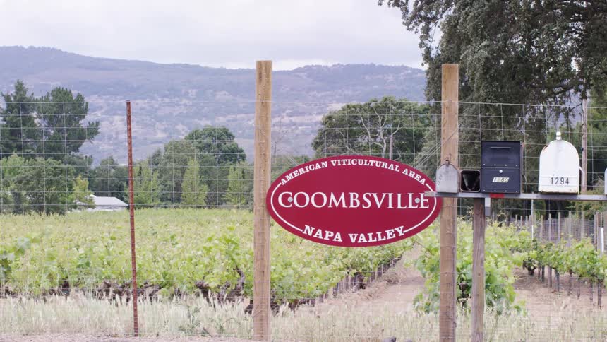 A red oval sign reading coombsville napa valley american viticultural area hangs on a fence overlooking a vineyard with hazy mountains in the background