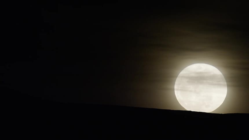 A large bright full moon partially obscured by a dark foreground a hill against a dark hazy sky