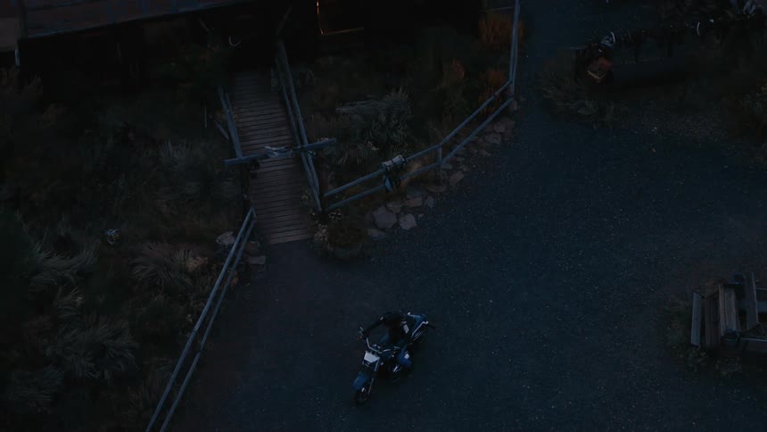From above a person mounts a motorcycle resting on a dark gravel path near a wooden staircase and sparse vegetation then rides away