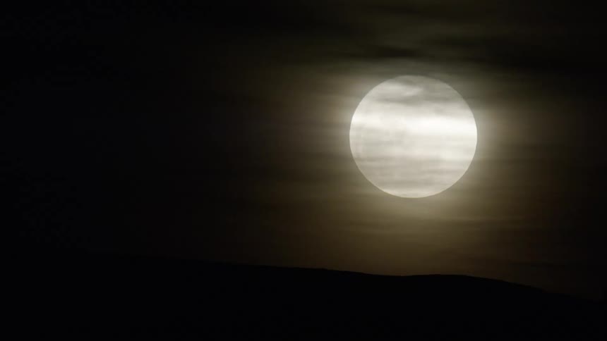 A luminous full moon its surface slightly obscured by wisps of clouds glows against a dark night sky