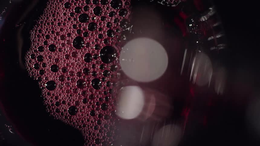 Red wine being poured into a glass forming a dense layer of bubbles against a dark background with blurred bokeh
