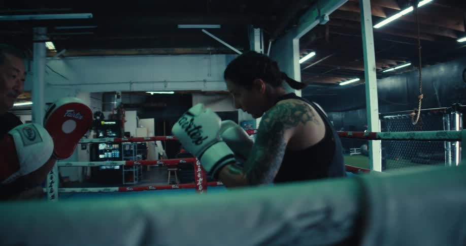 Inside a boxing gym a woman wearing boxing gloves and pads is training with a coach holding a padded target