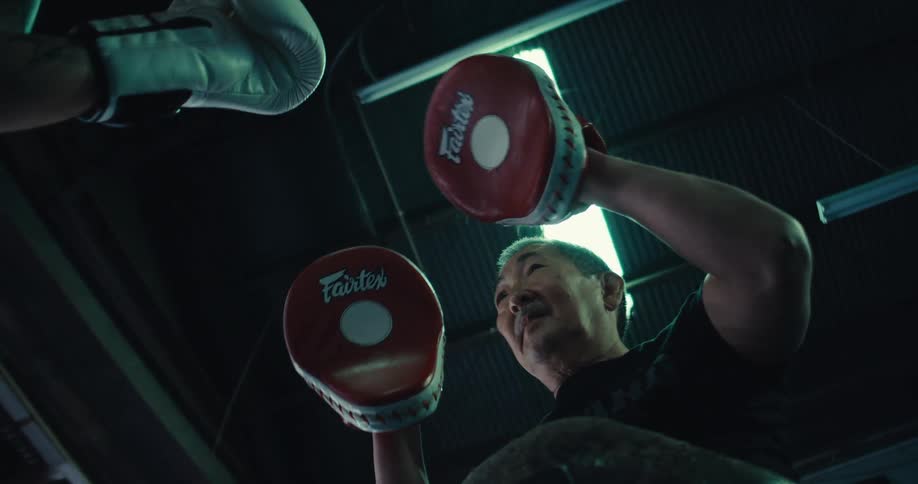 An older asian man has boxing pads on his hands and is helping someone off camera spar framed by a rectangular light over his head