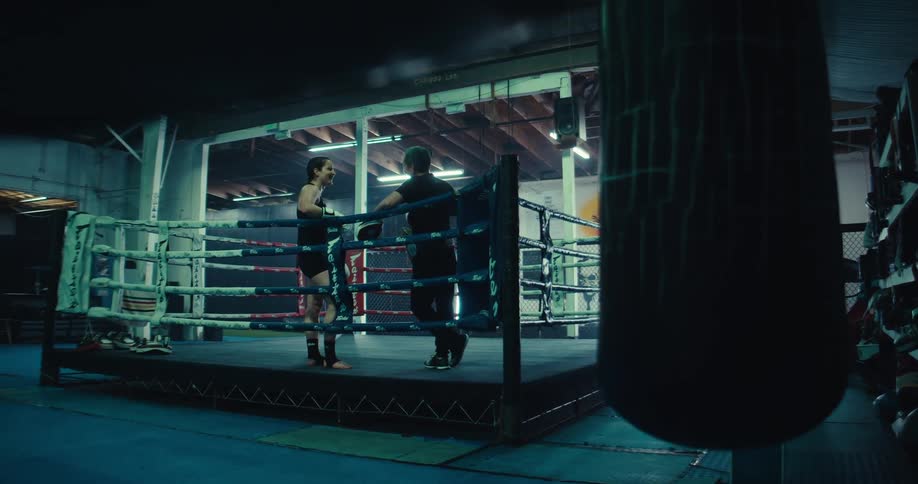 Inside a boxing gym two individuals stand in a boxing ring one appearing to be receiving instruction from the other