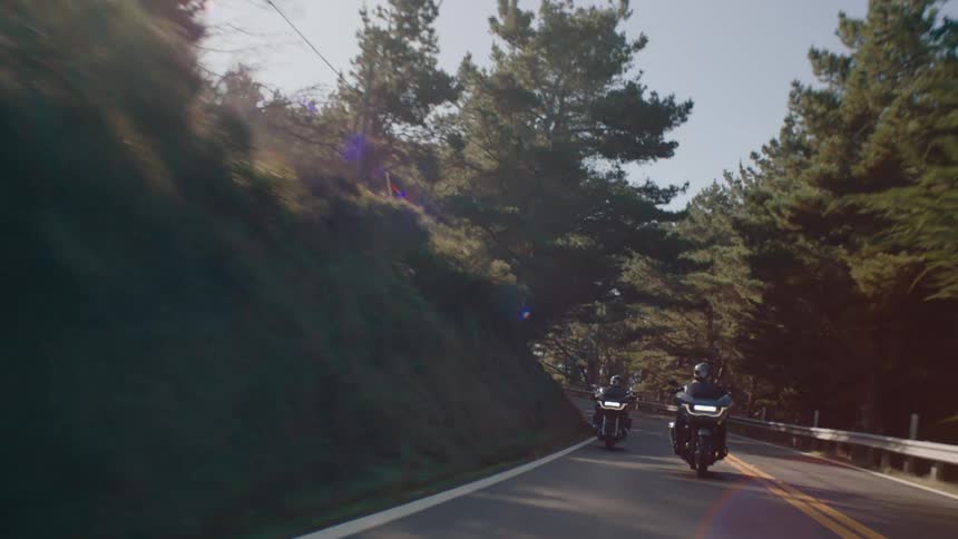 Two motorcyclists wearing dark riding gear and helmets riding in formation on a paved road that curves through a landscape featuring a steep dark hillside on the left and a sunlit area with trees on the right under a clear bright sky