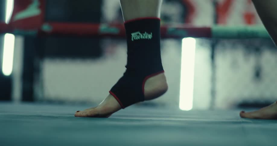 A closeup shot follows the moving bare feet of a kickboxer wearing black ankle supports as they perform footwork drills on a gym mat