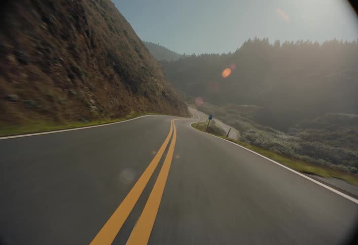 A moving vehicle captures an asphalt road curving along a coastal cliff with a metal railing separating the road from the ocean visible below all under a clear blue sky