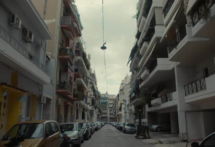 An urban street scene captures a narrow alleyway lined with tall apartment buildings on either side with parked cars stretching into the distance and a single hanging light fixture suspended above the street