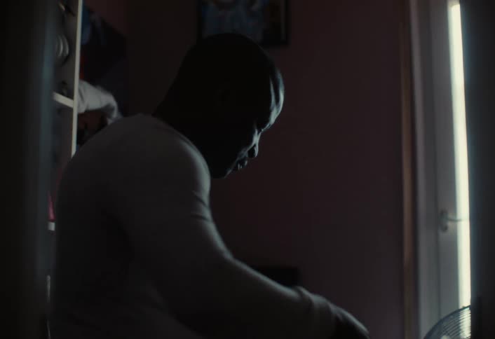 Man ironing his shirt on an ironing board in darken room near a window