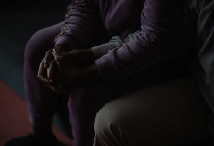 Two individuals sit closely together in a dimly lit setting holding each others hands