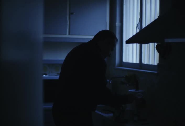 A silhouetted figure stands in a dimly lit kitchen area near a window with blinds