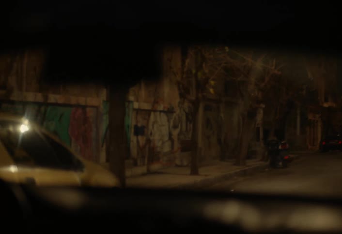 Through a vehicles windshield at night a dimly lit street scene with parked cars and buildings is visible