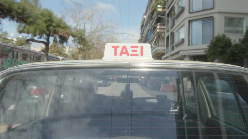 The rear window of a taxi with a sign on top reflects a street scene with buildings and trees under a partly cloudy sky