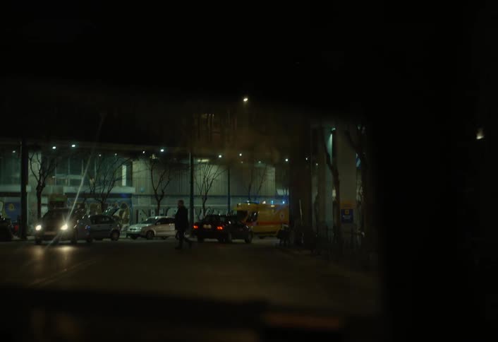 A view of city nightlife from a moving car shows an ambulance with flashing blue lights parked on a city street near a graffiticovered building