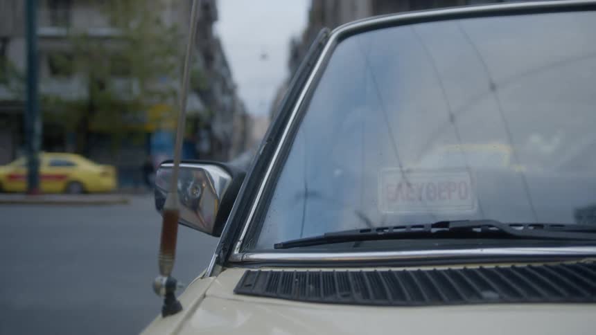 Front of a beige car with an antenna and a sign in greek on the windshield driving on a street