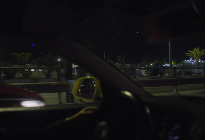 A drivers hands are on the steering wheel with blurred city lights and streetlights passing by outside the windshield and side mirror