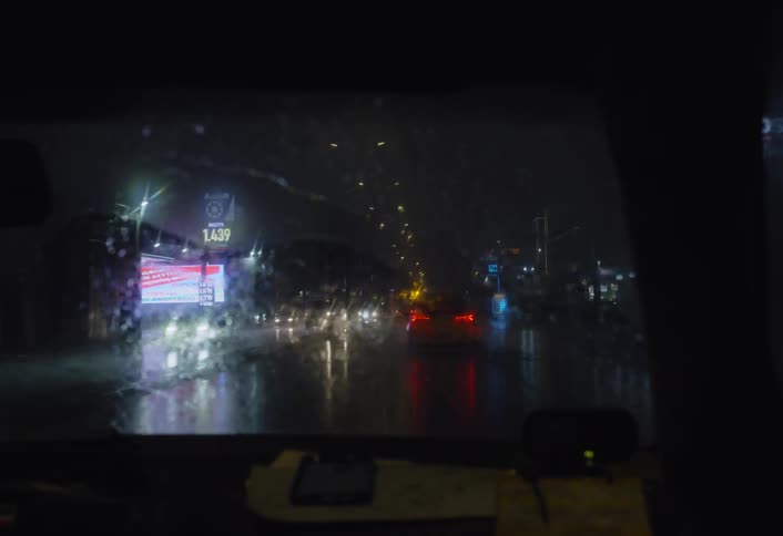 A mans silhouette is visible inside a dimly lit car at night with rain or moisture on the window and blurred lights in the background