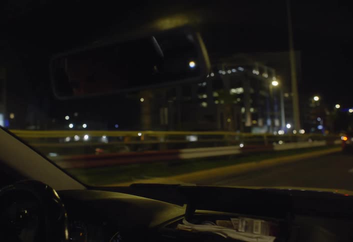 From inside a vehicle at night the glow of illuminated city buildings and the streaks of moving car lights are seen through the windows and windshield