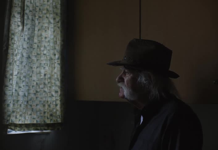 Older man wearing a black hat looking out window from dark room smoking a cigarette
