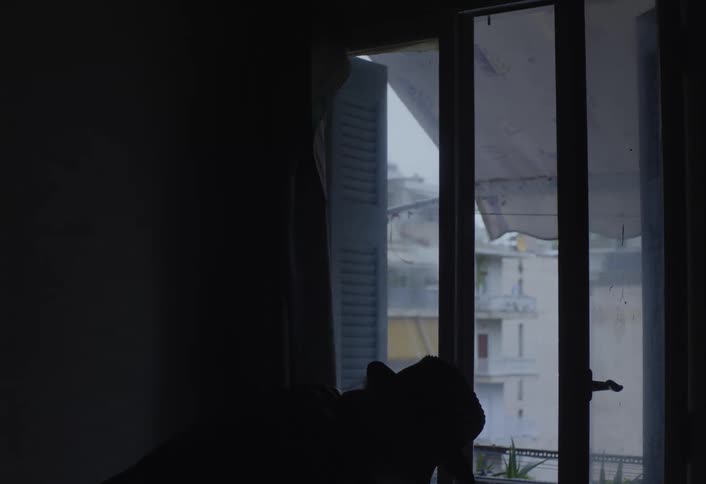 An older man wearing a long coat and hat opens a balcony door and steps outside from a dimly lit room revealing a shadowy silhouette against the light of surrounding city buildings