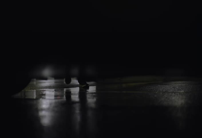 A persons foot is visible walking on wet asphalt viewed from beneath a parked vehicle at night