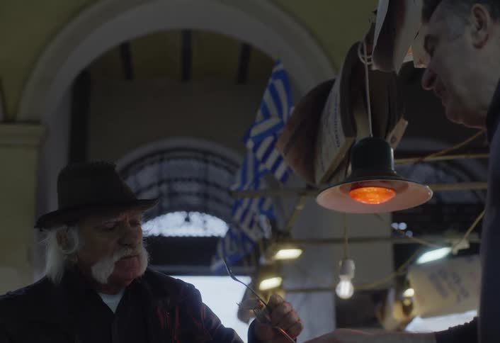 A man with a hat white hair and a large mustache samples food from a street vendor in a covered market adorned with greek flags