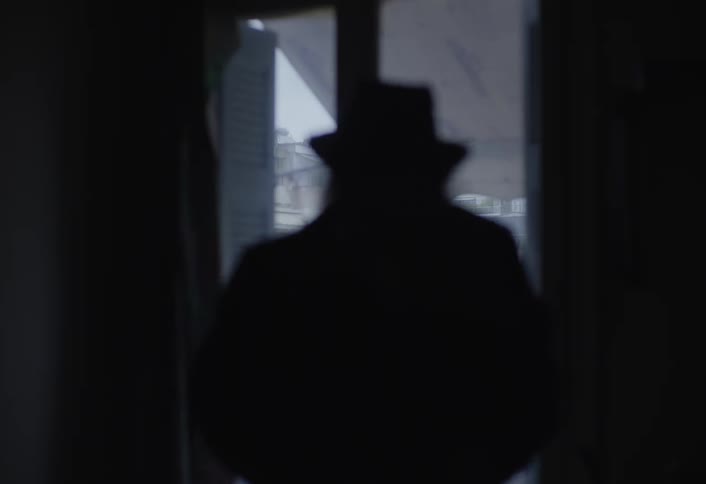 A silhouetted figure with long hair wearing a hat stands in a dimly lit room they are closing a glass door that reveals a bright overcast cityscape beyond