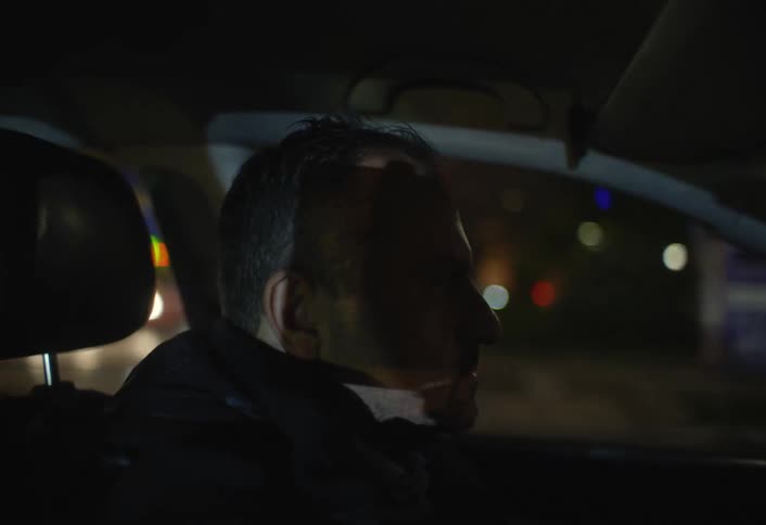 A mans silhouette is seen in the drivers seat of a car at night with the city lights blurred in the background