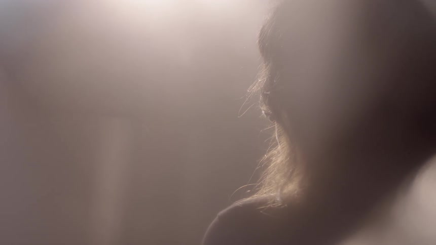 A woman standing in a room in sunlight raises her head to look at a window above her