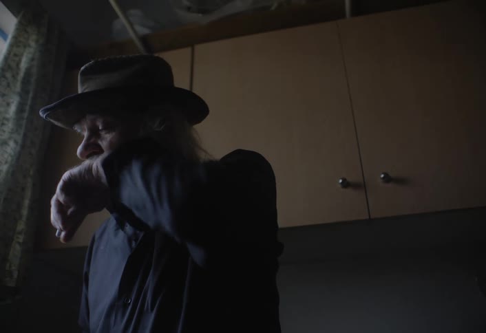 An elderly man with a flowing white beard wearing a dark hat sips from a tall glass through a straw while holding a lit cigarette