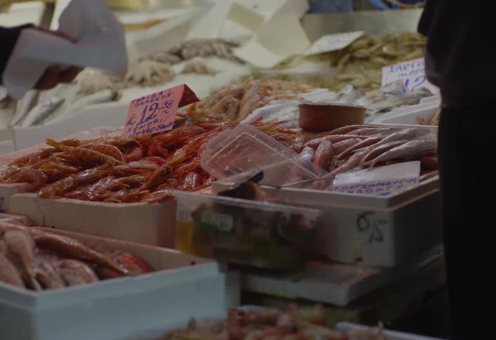 Various types of seafood are displayed in containers with price tags at what appears to be a bustling market stall