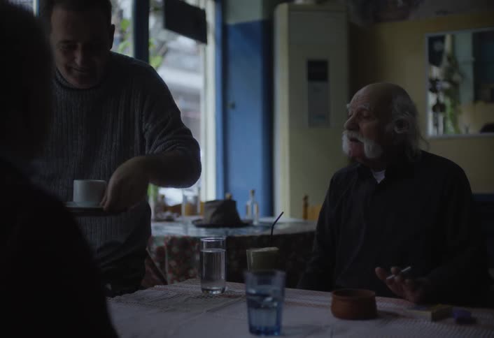 Inside a cafe a person serves a cup of coffee to another person seated at a table