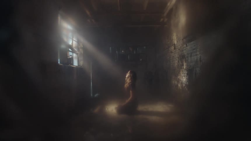 A woman is kneeling in a dark room as light peeks in through the window