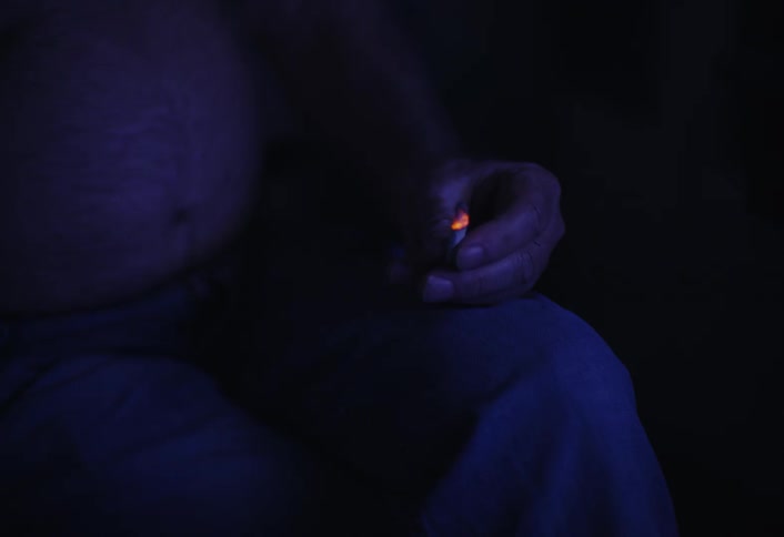 A closeup shows a mans belly as he sits at home smoking and changing tv channels with a remote in a dimly lit room