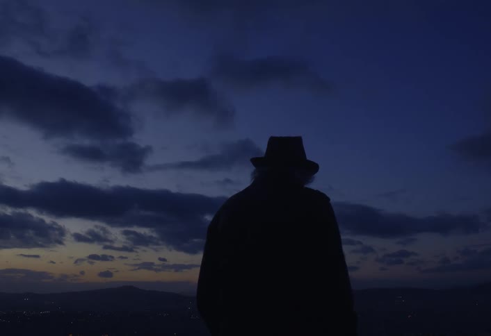 A silhouetted man wearing a hat stands against a twilight sky with clouds and a hint of a distant landscape visible in the background