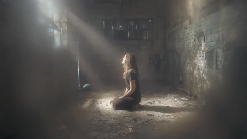 A woman is kneeling in front of a window giving off light in a dark room