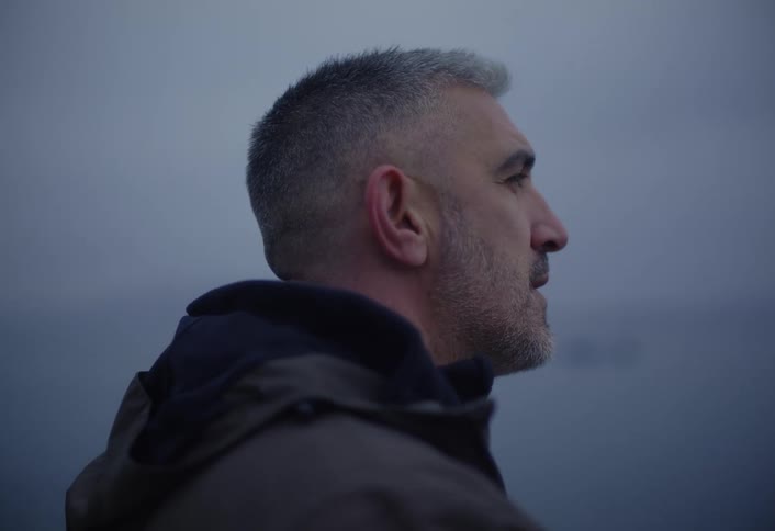 A man with short gray hair wearing a dark jacket is seen in profile smoking a cigarette against a foggy or overcast background by the sea