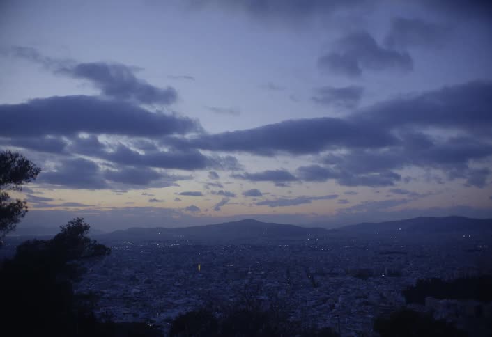A still mountaintop view overlooks a sprawling city as dusk settles in casting soft orange and purple hues across the cloudfilled sky
