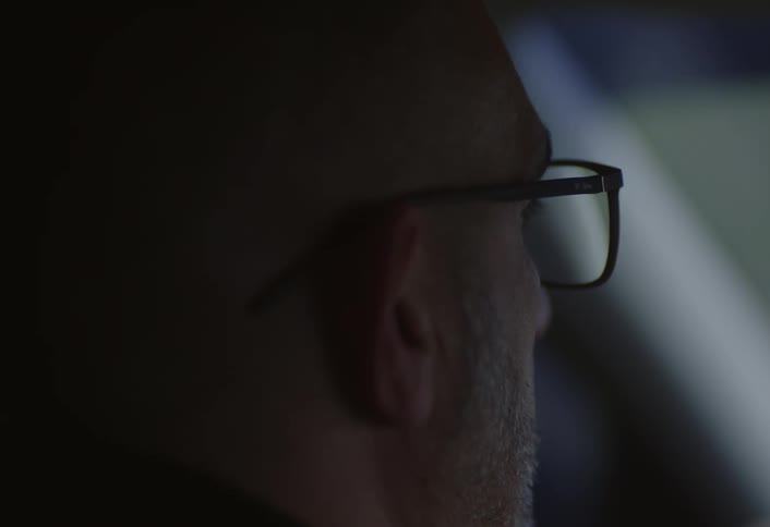 The side profile of a mans face wearing glasses against a dark shadowy background