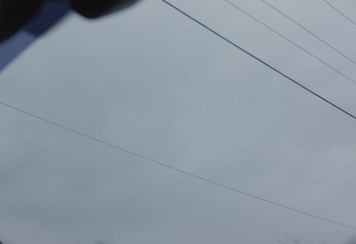 Through the windshield of a moving vehicle with wipers in motion a blurred scene of power lines against an overcast sky comes into view