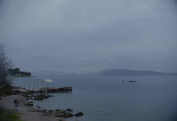 A person walks near a calm body of water that meets a rocky shore with a pier under a gray sky revealing a distant land