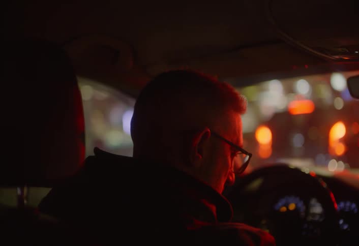 From behind a person wearing glasses is seen driving a car at night with blurred city lights visible through the windshield