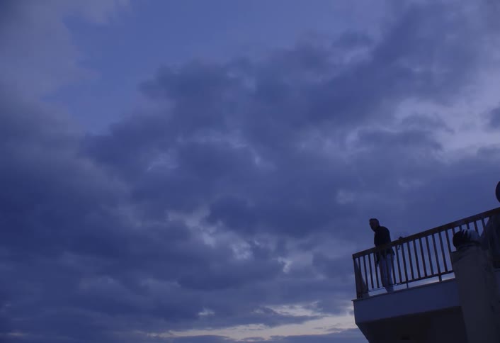 A silhouetted figure stands on a railed platform under a cloudy twilight sky