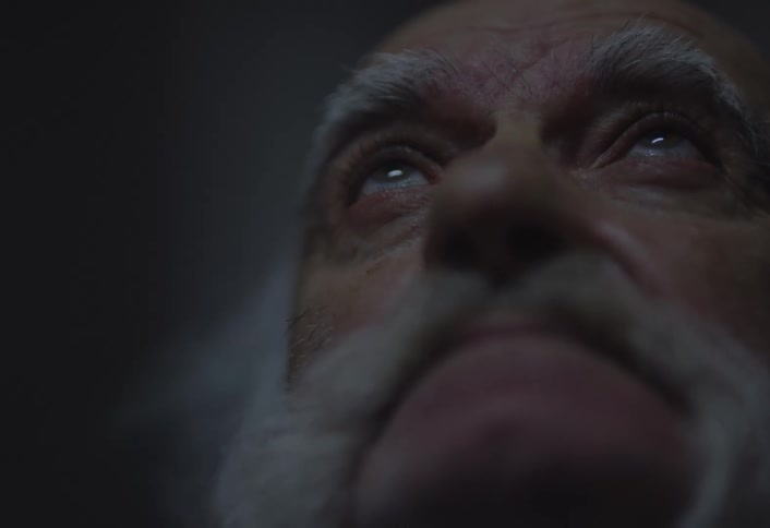 A closeup lowangle shot shows the face of an older individual with a white beard and eyebrows looking upwards with lightcolored eyes