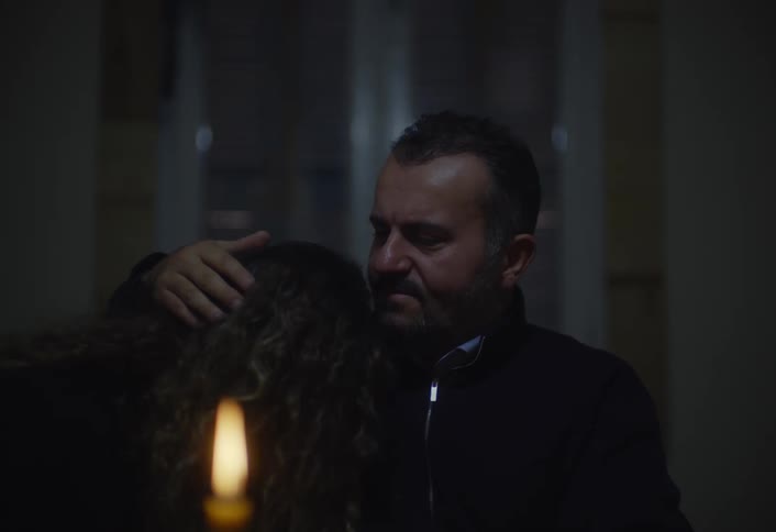 A man sits on a chair consoling a woman whose head rests on his chest gently stroking her hair in a dimly lit room illuminated by a candle