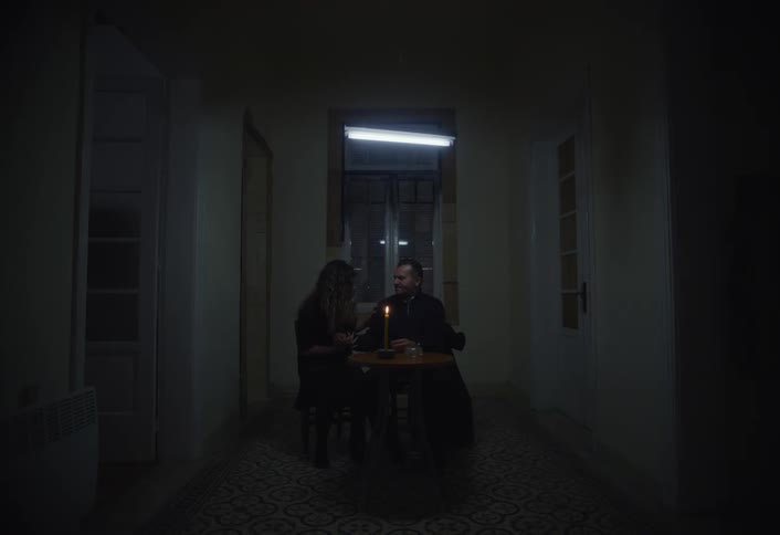 In a dimly lit hallway a man and a woman sit at a small table illuminated by a single candle engaging in conversation