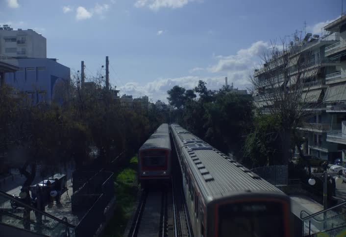 Two trains pass each other on parallel tracks beneath a pedestrian bridge while people walk up the steps on the left side in a sunny urban neighborhood